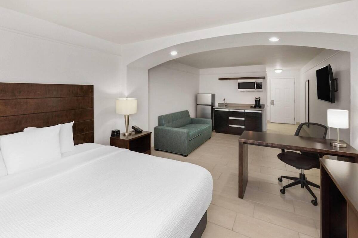 A hotel room with a bed, a desk, a chair, a sofa, a kitchenette, and a TV.