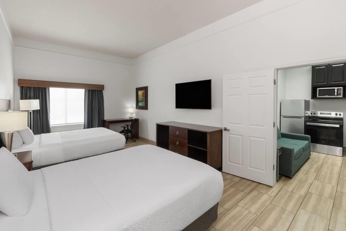 A hotel room with two beds, a desk, a TV, a kitchenette, and a sliding door to the bathroom.