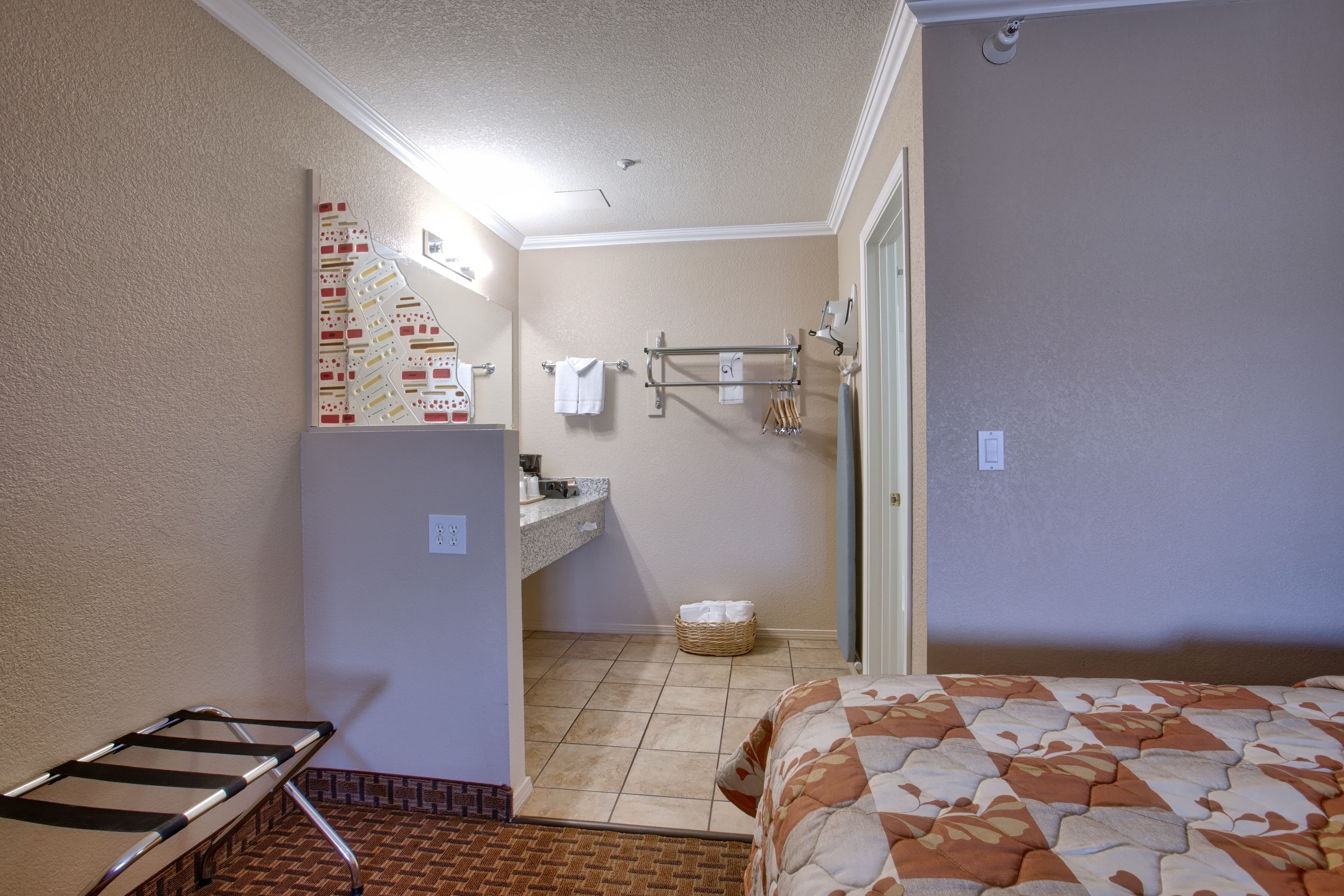 A compact hotel room with a bed, a small bathroom with a sink and towel rack, and a laundry basket.