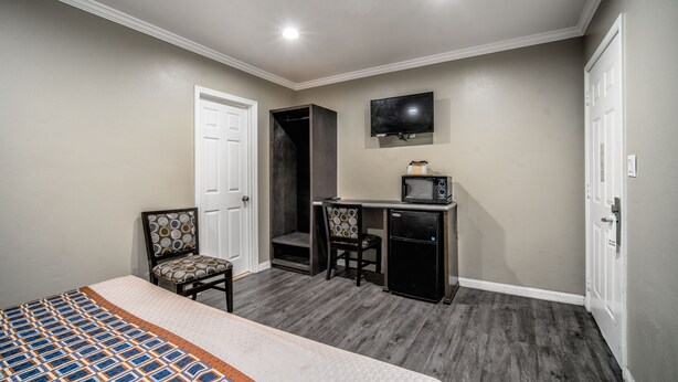 A bedroom with a bed, a desk with a TV, a chair, and a small refrigerator. - Regency Inn & Suites (Downey)