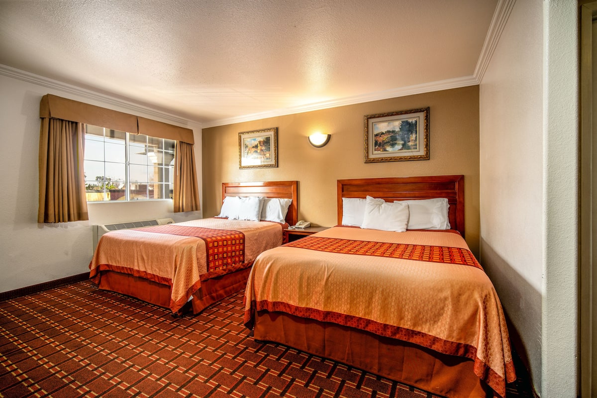 Une chambre d’hôtel avec deux lits, une fenêtre avec des rideaux, un téléphone fixé au mur et deux tableaux encadrés.