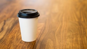 A white disposable coffee cup with a black lid on a wooden surface. - Regency Inn & Suites (Downey)