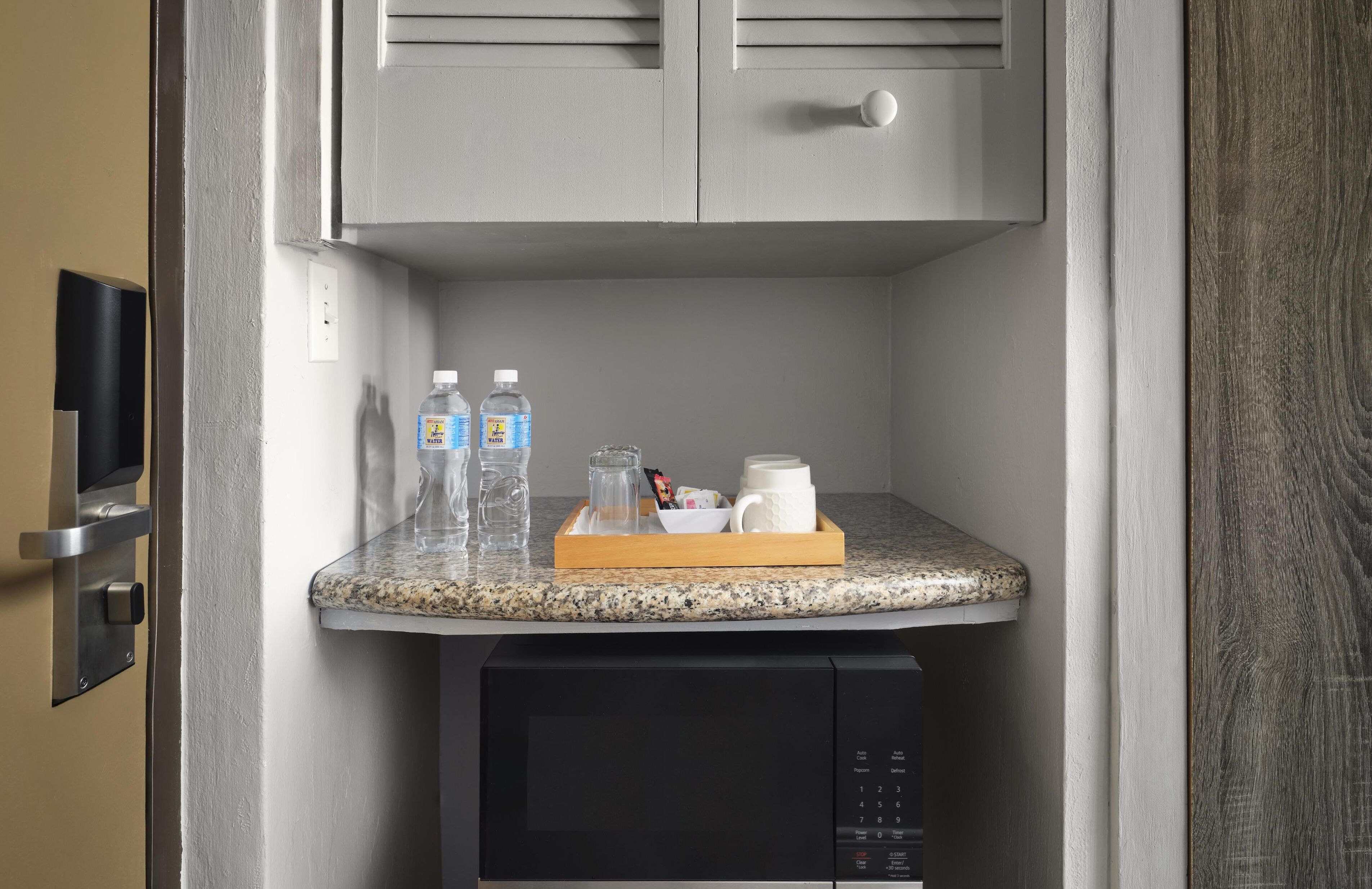 A compact kitchen area with a granite countertop, a microwave, and a small refrigerator. There are two bottles of water and a coffee maker on the counter.