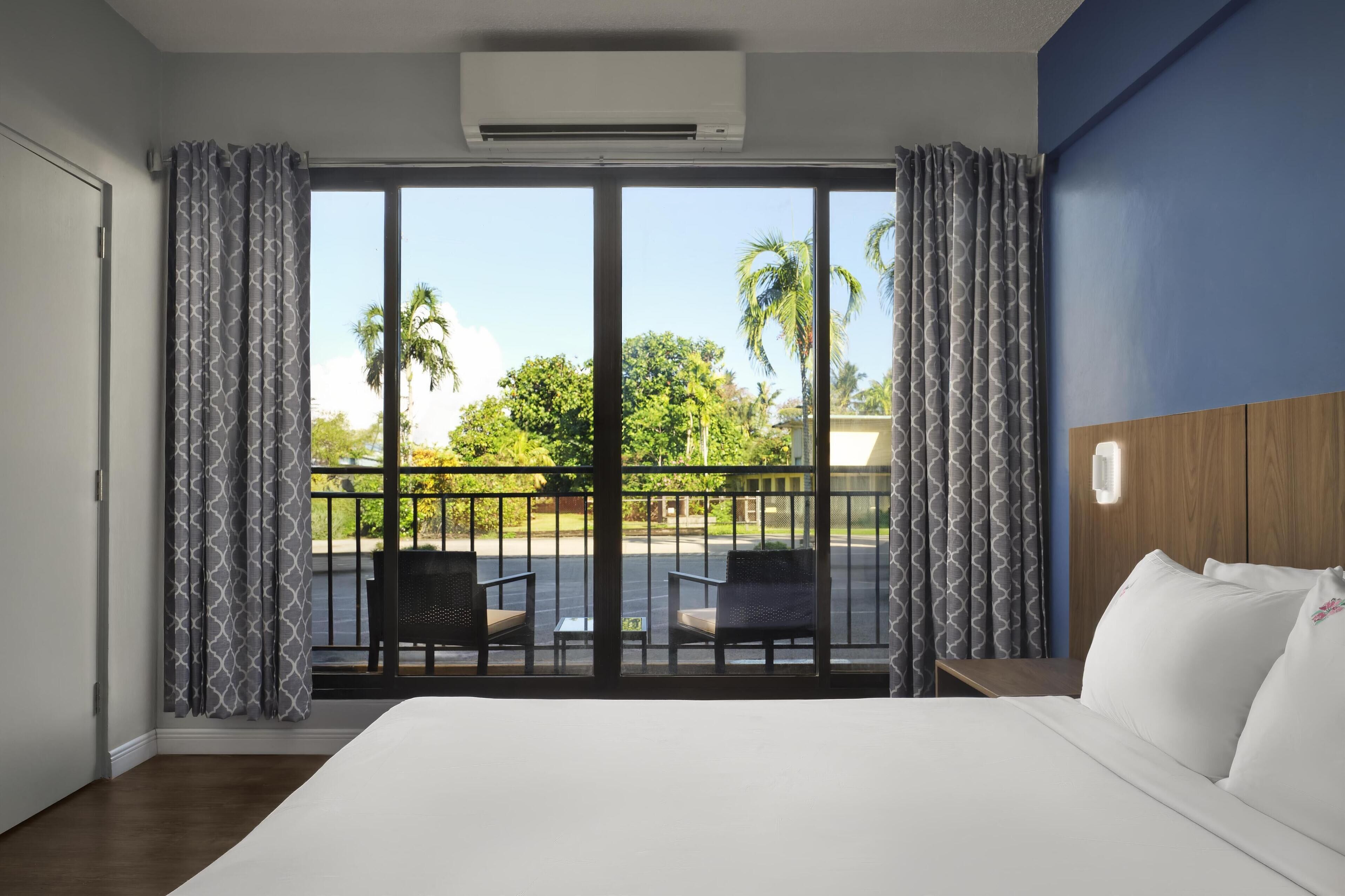 A hotel room with a large bed, a balcony with chairs, and a view of palm trees.