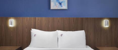 A hotel room with a wooden headboard, white bedding, and a framed abstract painting on the wall.