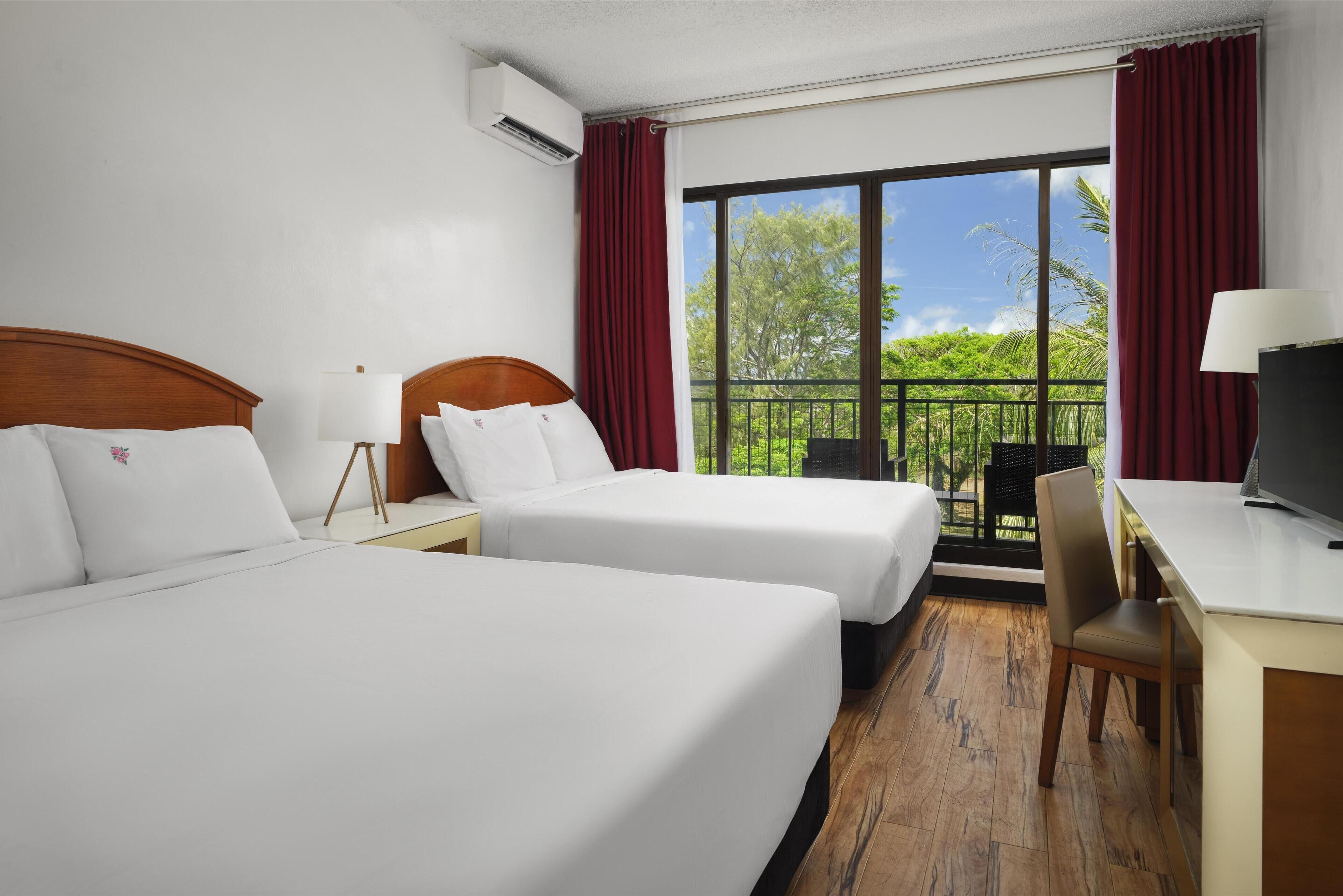 A hotel room with two beds, a desk, a chair, and a large window with a balcony view.