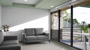 A modern waiting area with a grey sofa, a white chair, and a large window with a view of a stone wall and greenery. - SureStay Hotel by Best Western Guam Airport South (Barrigada)