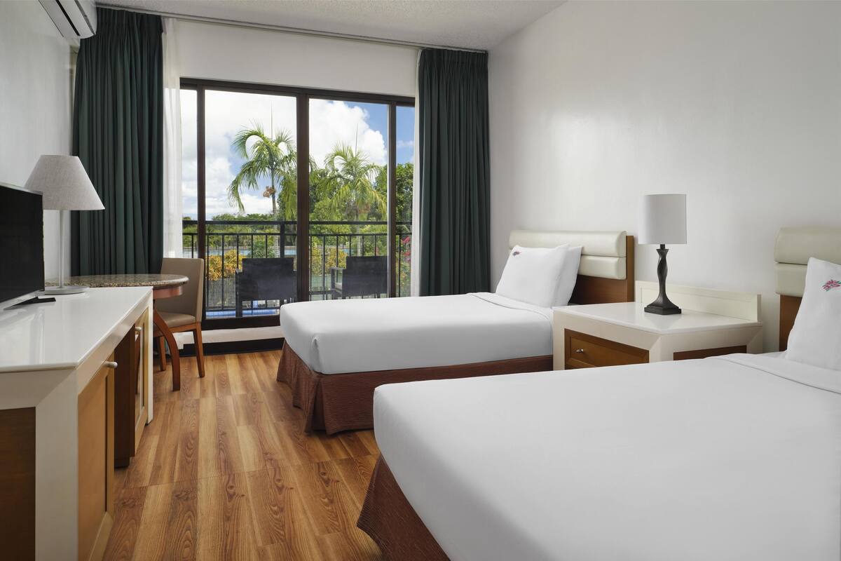 Ein Hotelzimmer mit zwei Betten, einem Schreibtisch, einem Stuhl und einem Balkon mit Blick auf Palmen.