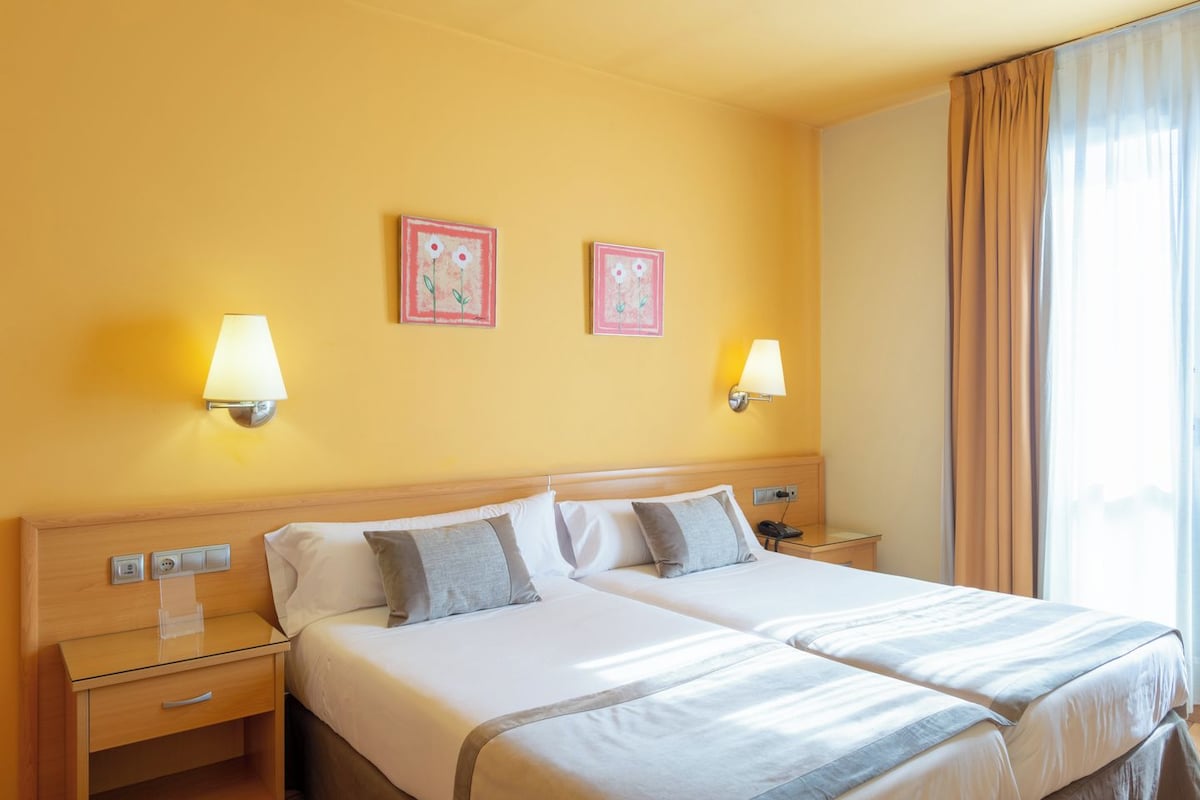 Una cama doble con cabecera de madera, dos mesitas de noche, dos lámparas de pared y dos cuadros enmarcados en la pared.