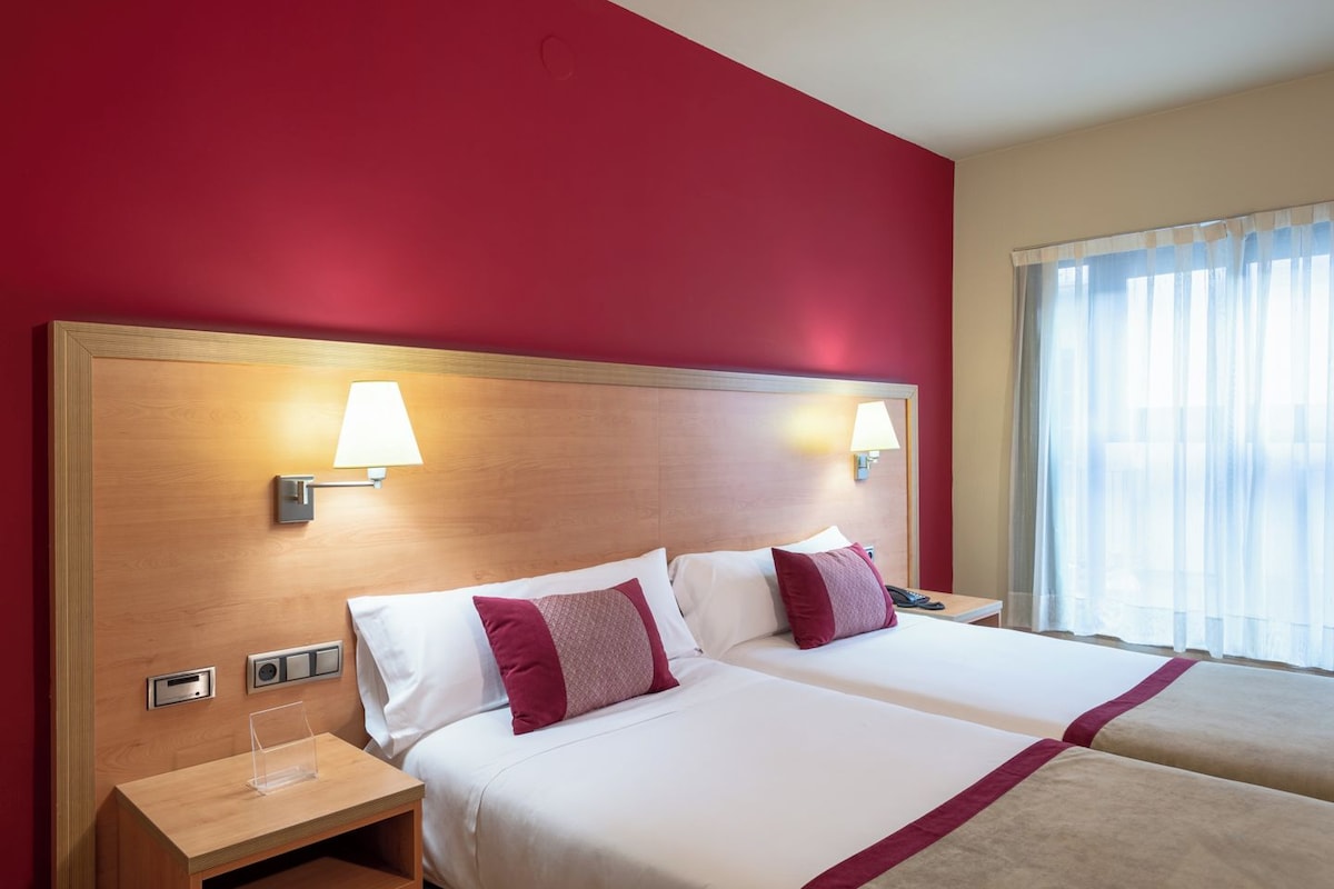 Une chambre d’hôtel avec deux lits, une tête de lit en bois, des lampes de chevet, une table de chevet et une fenêtre avec des rideaux légers.