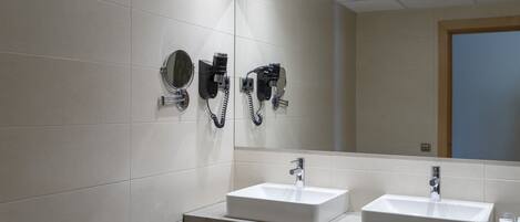 A modern bathroom with two sinks, a large mirror, and hair dryers mounted on the wall.