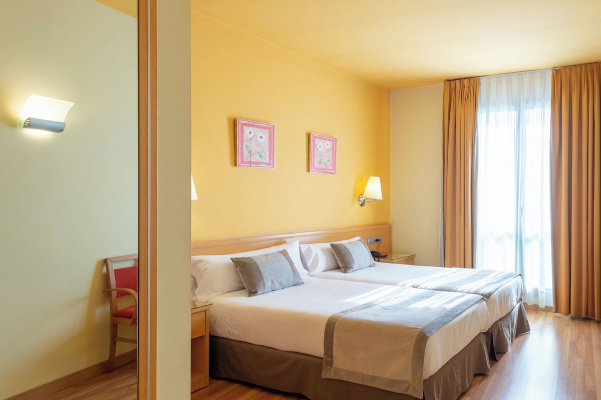 Habitación de hotel con dos camas, piso de madera, una mesita de noche, una lámpara y una ventana con cortinas.