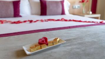 Una cama con una colcha blanca y morada, una decoración en forma de corazón rojo, y una bandeja blanca con cubos dorados.
