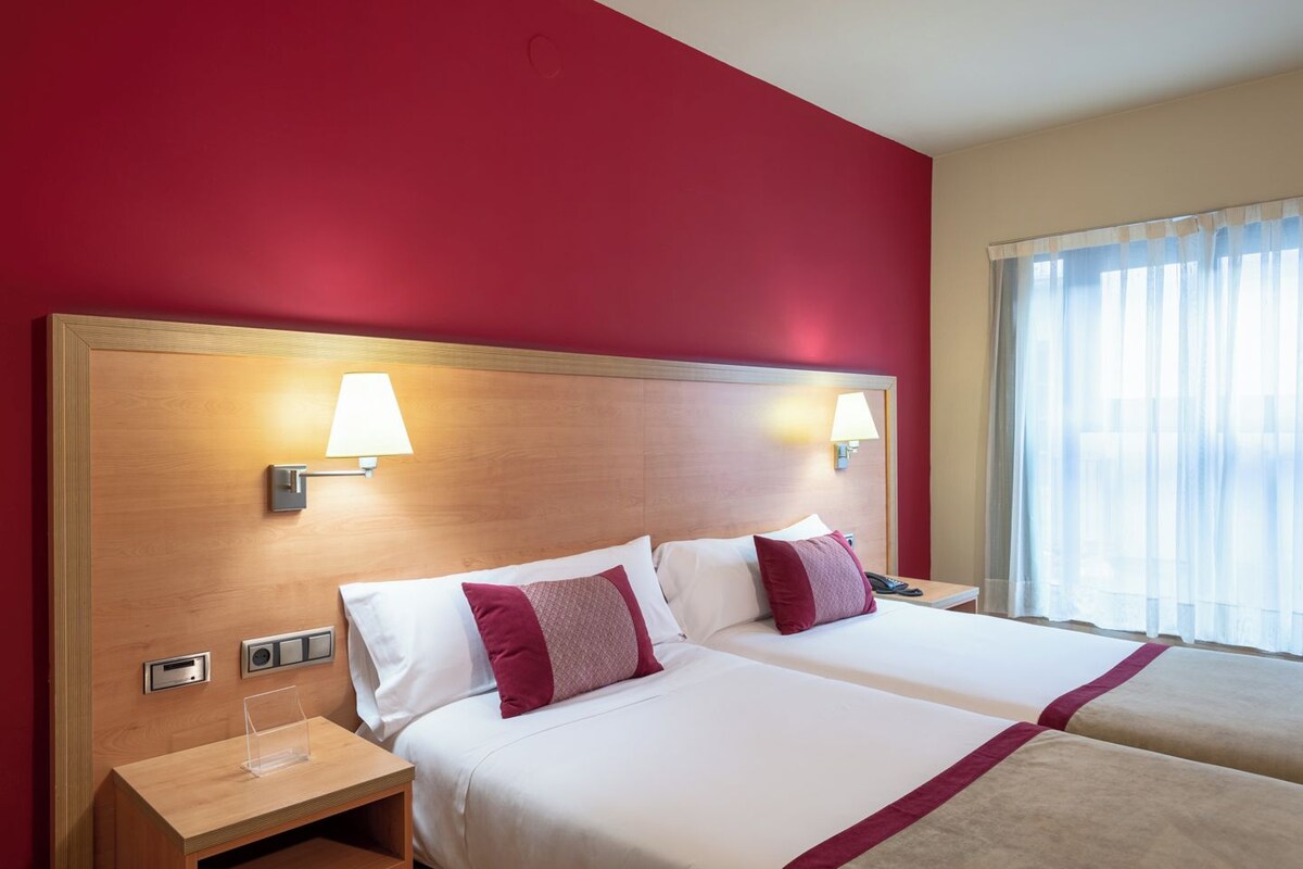A hotel room with two beds, a wooden headboard, bedside lamps, a nightstand, and a window with sheer curtains.