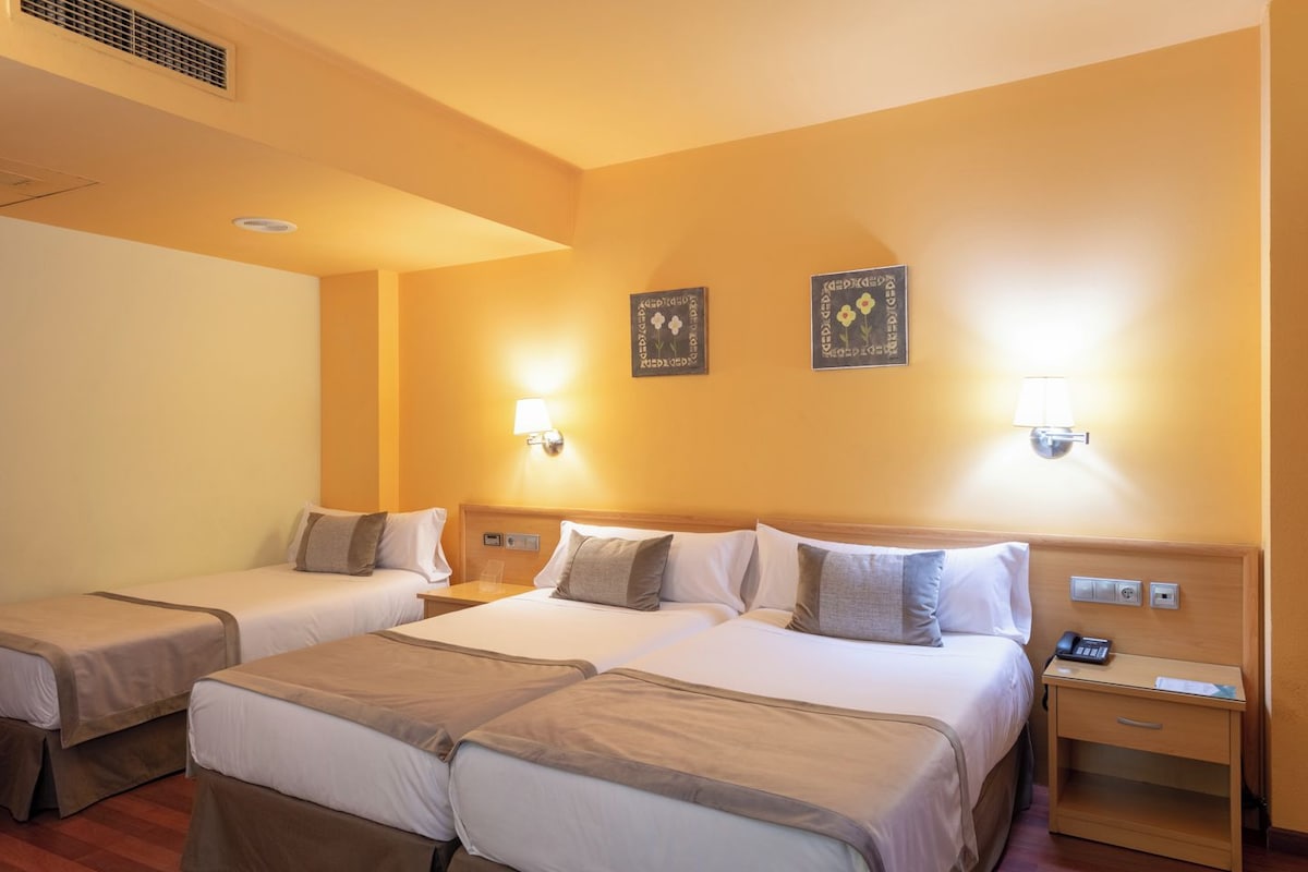 Habitación de hotel con dos camas, cada una con mesita de noche y lámpara. Se observan dos luces fijadas a la pared y una pequeña mesita con un teléfono.
