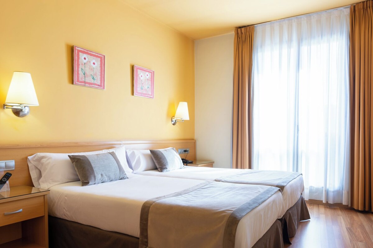 Habitación de hotel con dos camas, una mesita de noche con lámpara y una ventana con cortinas.