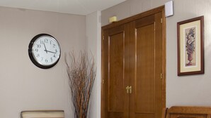 Una habitación con un armario de madera, un sillón a rayas, un banco, un reloj de pared y un florero con ramas.
