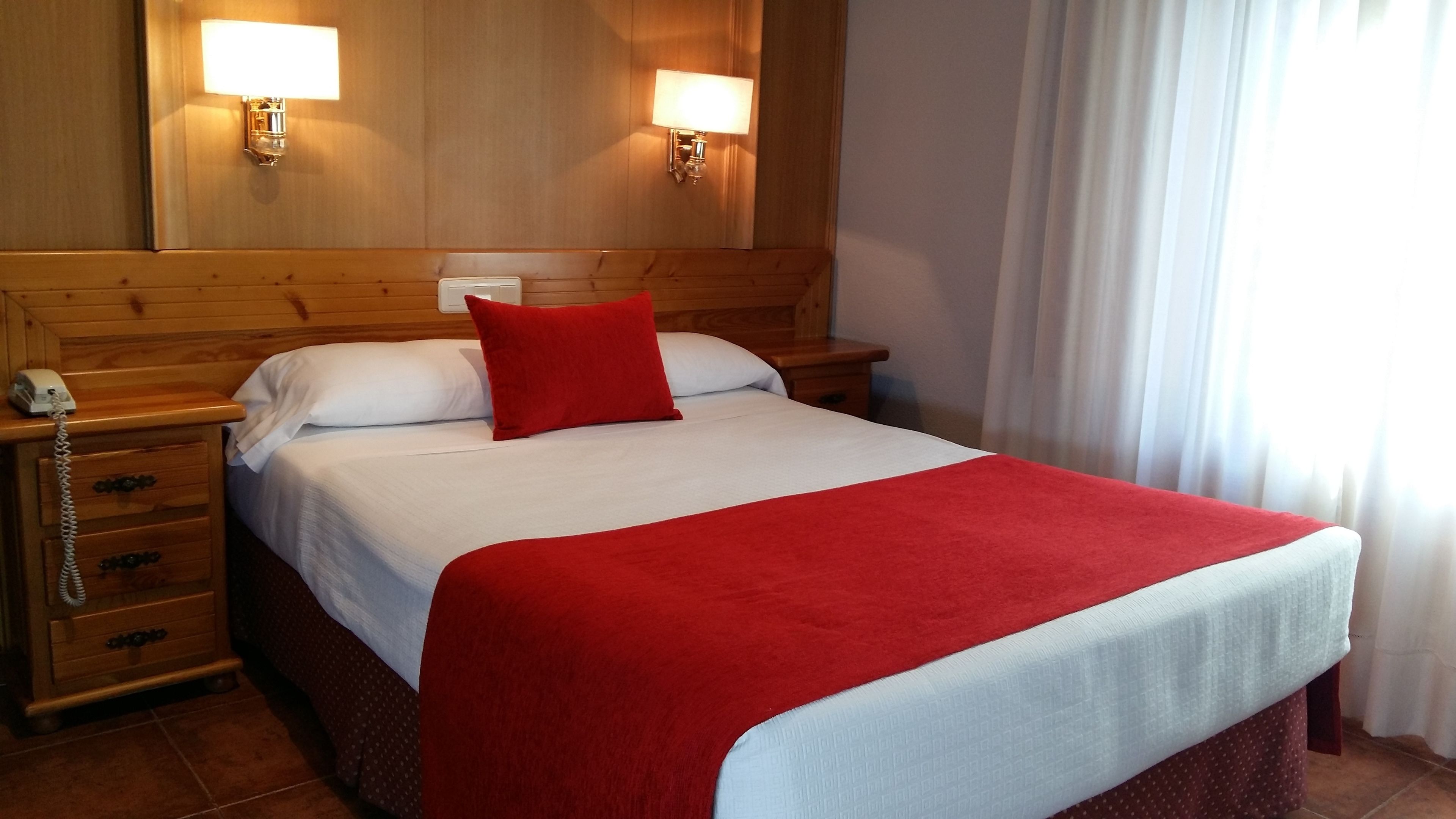 Una habitación de hotel con cabecera de madera, cama con ropa blanca y roja, dos lámparas de noche, un teléfono y una mesita de noche con cajones.