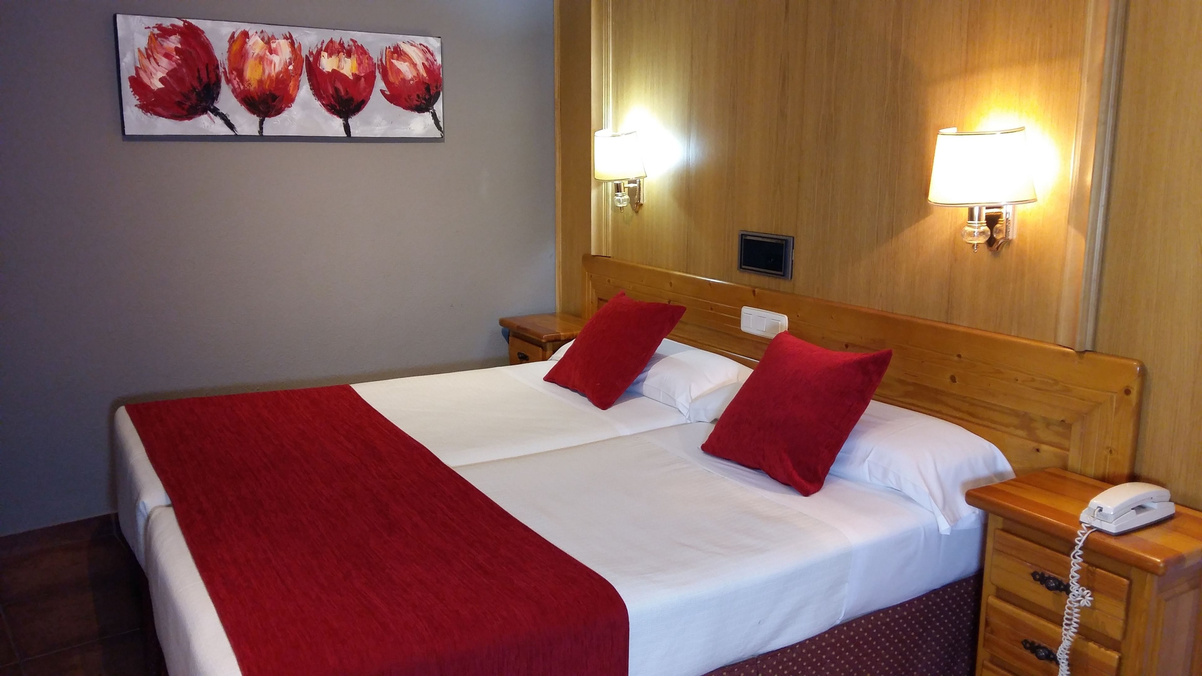 Habitación de hotel con una cama grande, dos almohadas rojas, una colcha blanca, un plaid rojo, un cabecero de madera, dos mesitas de noche con lámparas, un teléfono y un cuadro de flores en la pared.