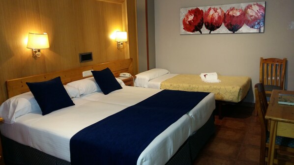 A hotel room with two beds, each with white linens and dark blue accents. There are wall-mounted lamps on either side of the beds, a painting of flowers on the wall, and a wooden chair and table in the corner.