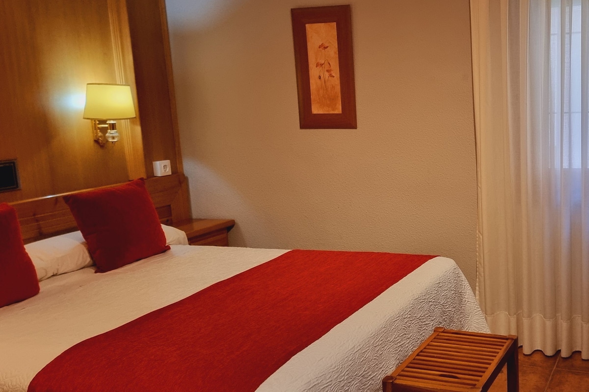 A hotel room with a bed, a wooden bench, a chair, a wall-mounted lamp, and a window with sheer curtains.