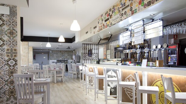 Un restaurante con sillas y mesas blancas, una zona de bar con grifos y botellas, y una pared decorada con varios logotipos.