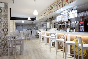 Un restaurante con sillas y mesas blancas, una zona de bar con grifos y botellas, y una pared decorada con varios logotipos.