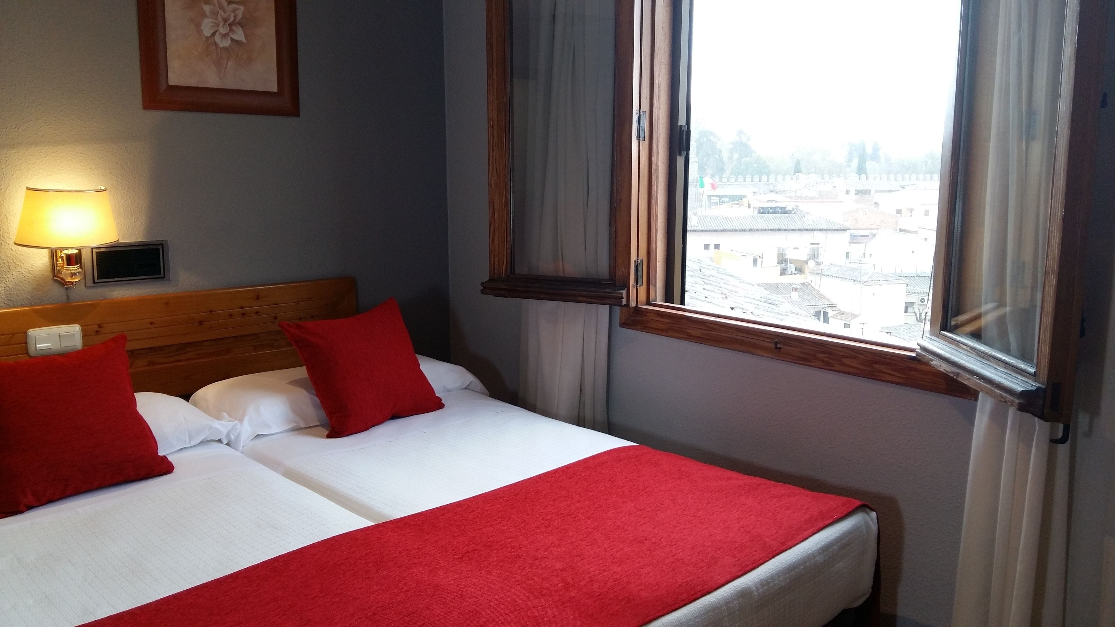 Habitación de hotel con cama, almohadas rojas, colcha blanca, manta roja, mesita de noche con lámpara y vista a edificios a través de una ventana.