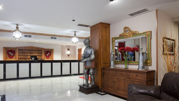 A lobby with a knight statue, a leather sofa, a wooden reception desk, and a mirror with a gold frame. - Hotel Real de Toledo (Toledo)