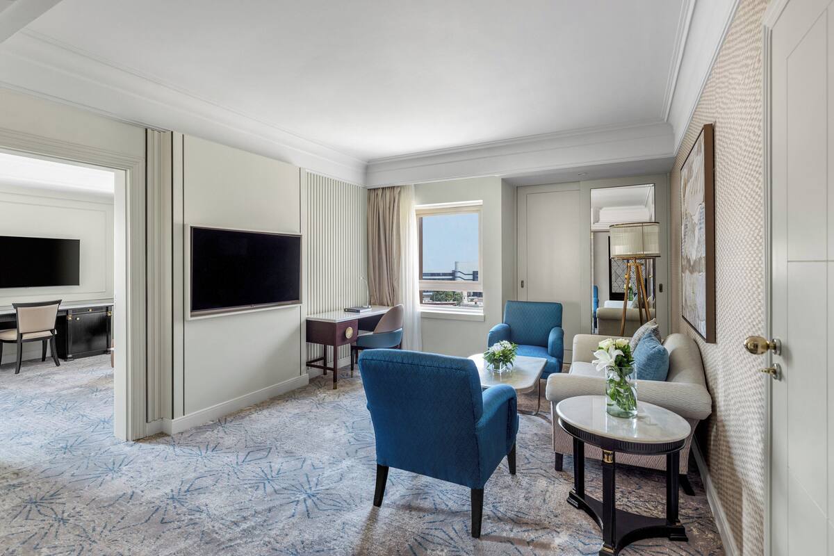 A hotel room with a large flat-screen TV, a sofa, a chair, a small table with a vase, and a view of the city through the window.