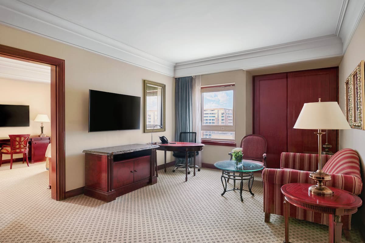 A hotel room with a desk, a TV, a sofa, a chair, a table, and a window with a view of buildings.