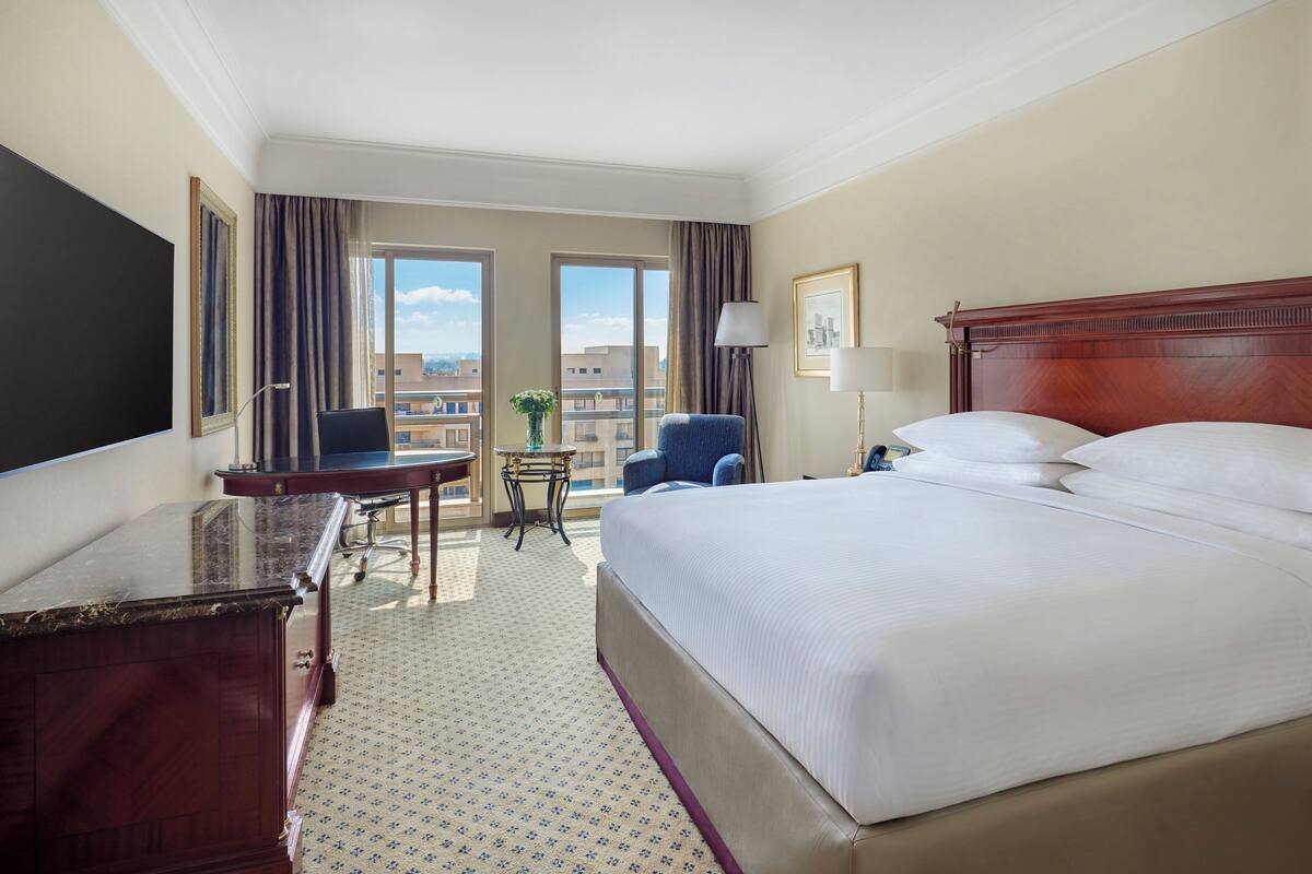 A hotel room with a large bed, a desk, a chair, a small table, and a balcony with a view of the city.