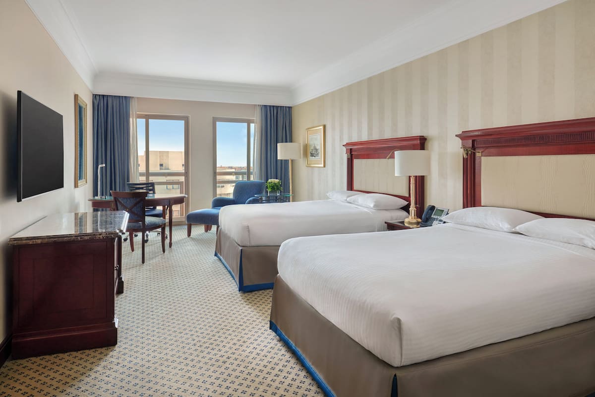 A hotel room with two beds, a desk, a chair, a TV, and a window with curtains.