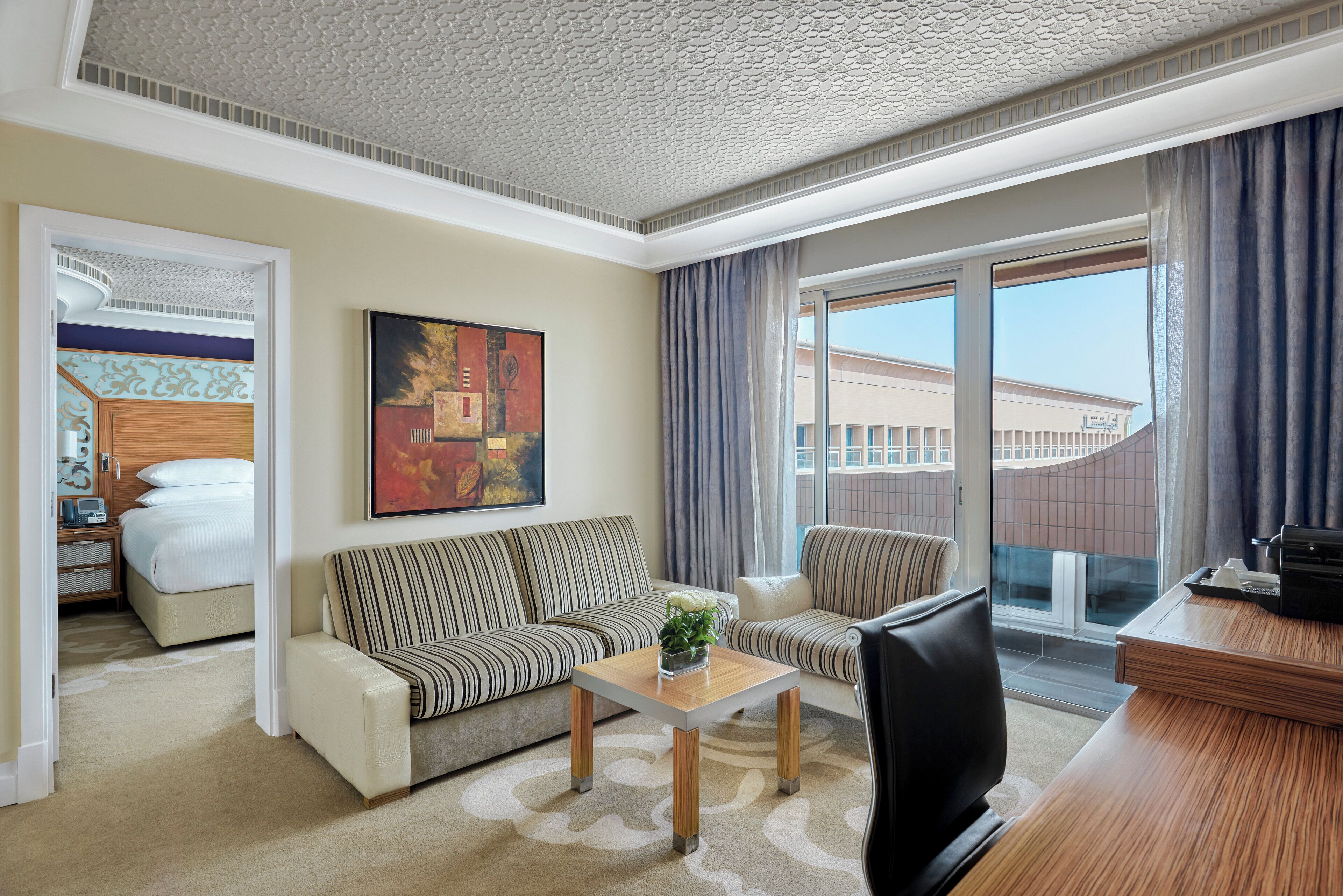 A hotel room with a sofa, a small table with a vase, a TV, and a desk with a computer. There is a window with curtains and a view of buildings outside.