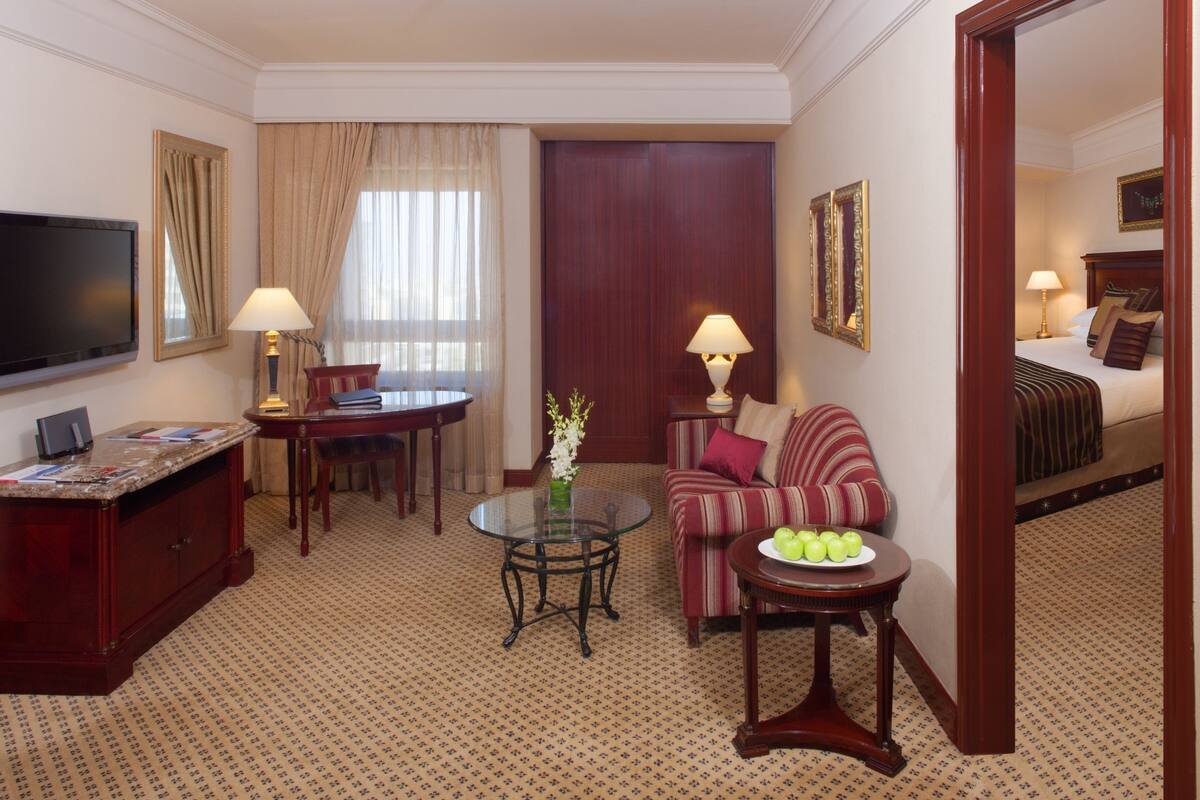 A hotel room with a TV, a dining table, a sofa, a small round table with apples, and a bed with pillows.