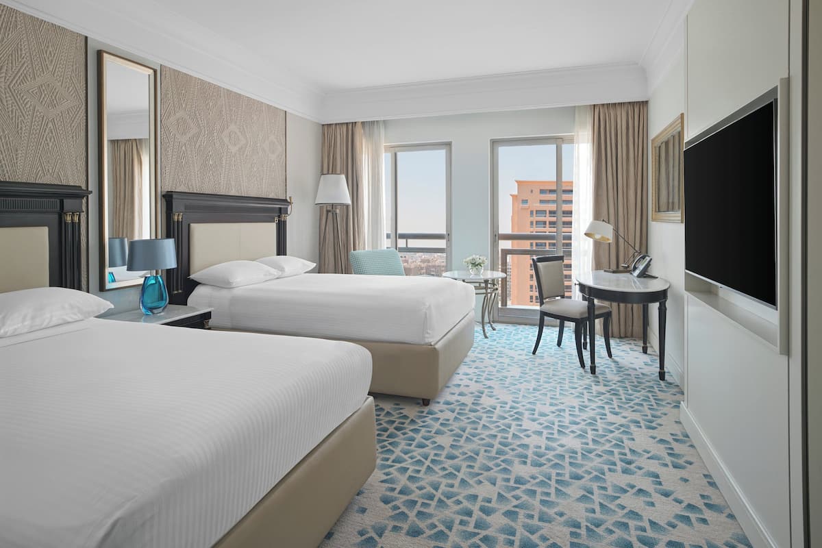 A hotel room with two beds, a desk, a chair, a TV, and a balcony with a city view.