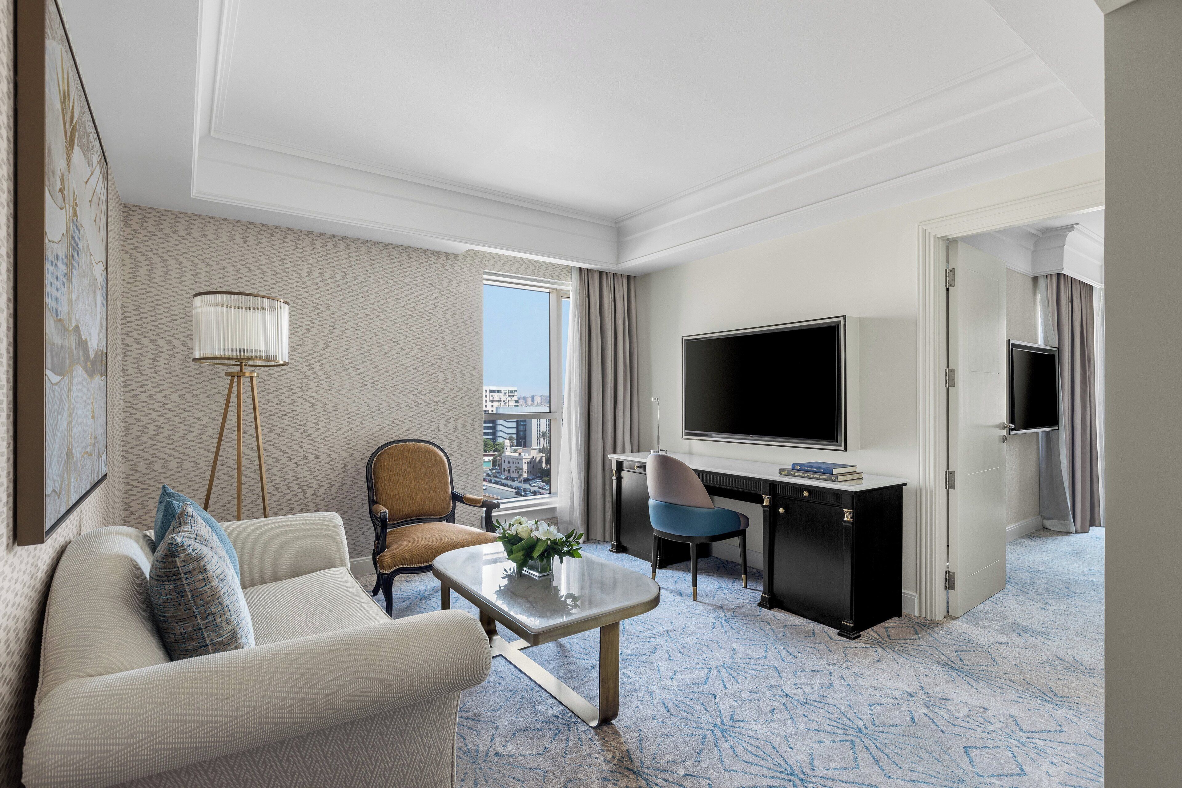 A hotel room with a sofa, armchair, desk, television, and a view of the city.