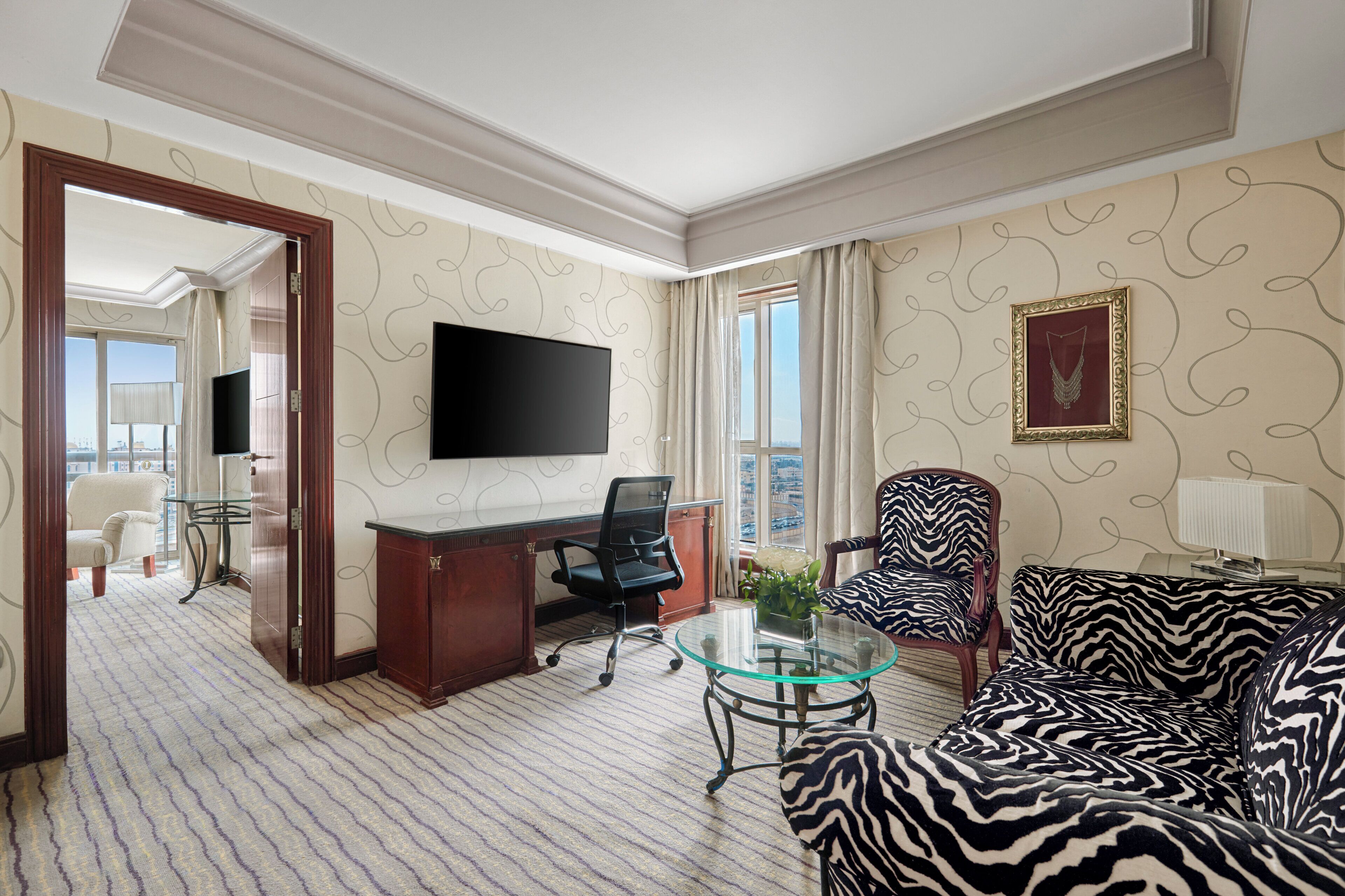 A hotel room with a large flat-screen TV, a zebra-patterned sofa, a glass coffee table, a desk with a chair, and a view of the city through the window.