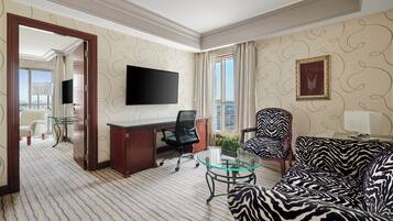 A hotel room with a large flat-screen TV, a zebra-patterned sofa, a glass coffee table, a desk with a chair, and a view of the city through the window.