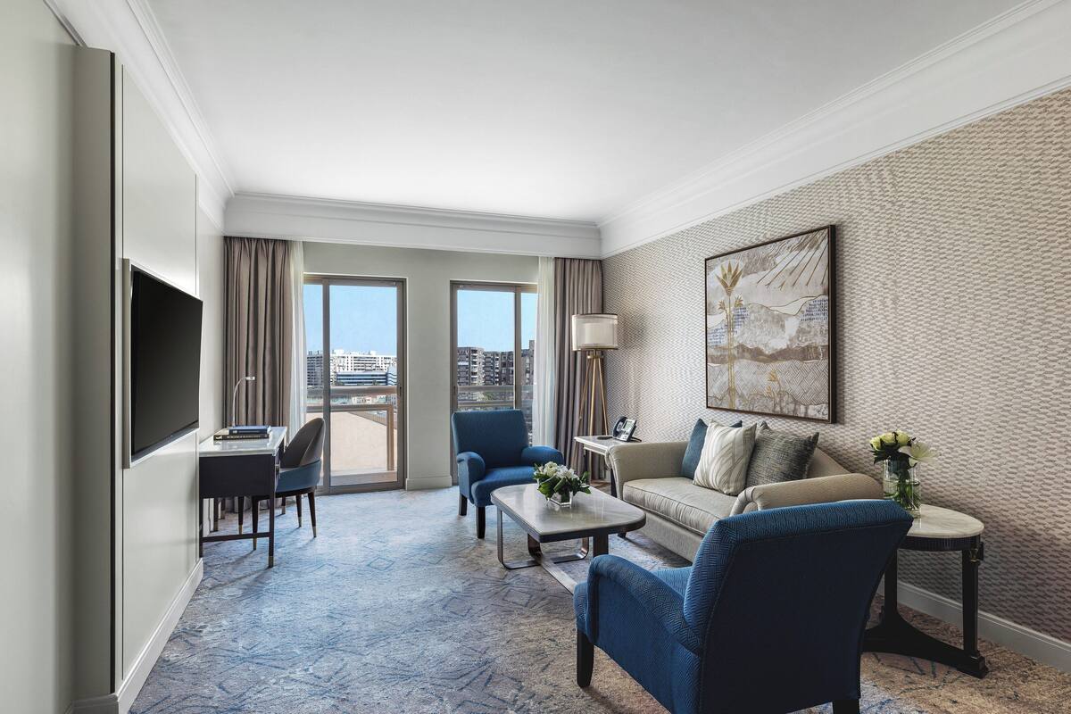 A hotel room with a flat-screen TV, a sofa, a chair, a coffee table, and a view of the city through large windows.