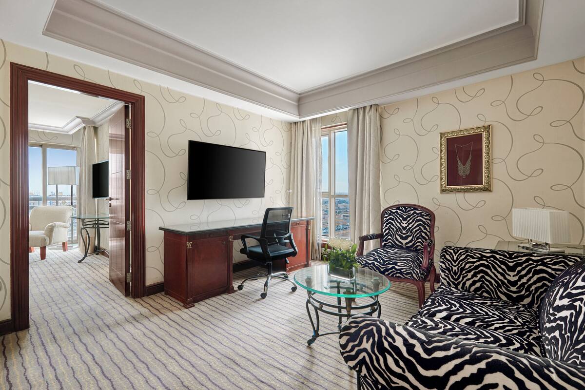 A hotel room with a large flat-screen TV, a zebra-patterned sofa, a glass coffee table, a desk with a chair, and a view of the city through the window.