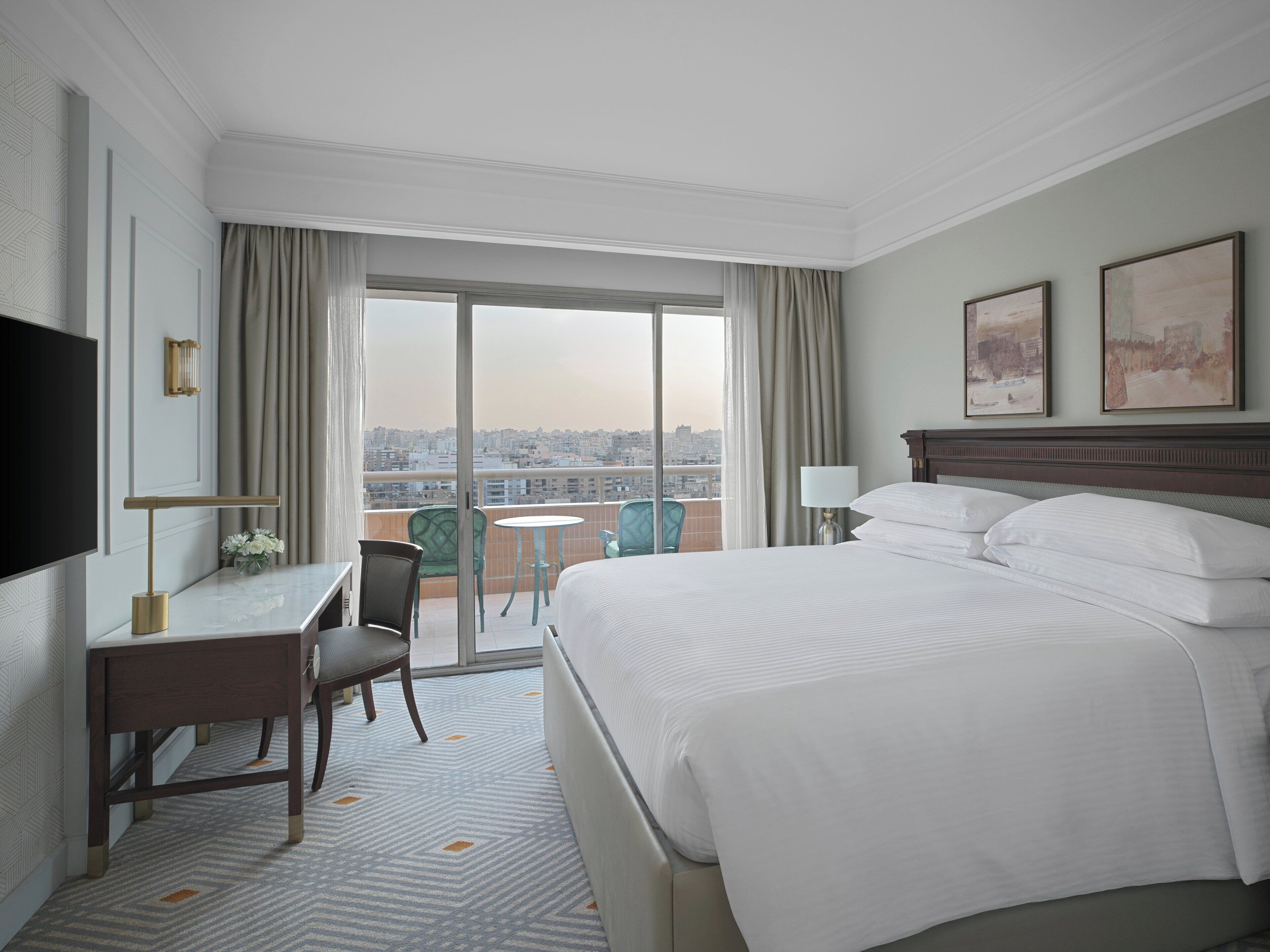 A hotel room with a large bed, a desk, a chair, a TV, and a balcony with a city view.