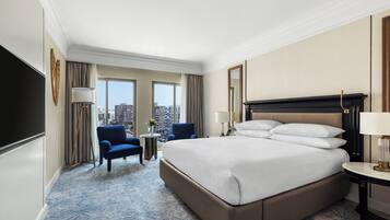 A hotel room with a large bed, a blue armchair, a small table, and a city view through the window.