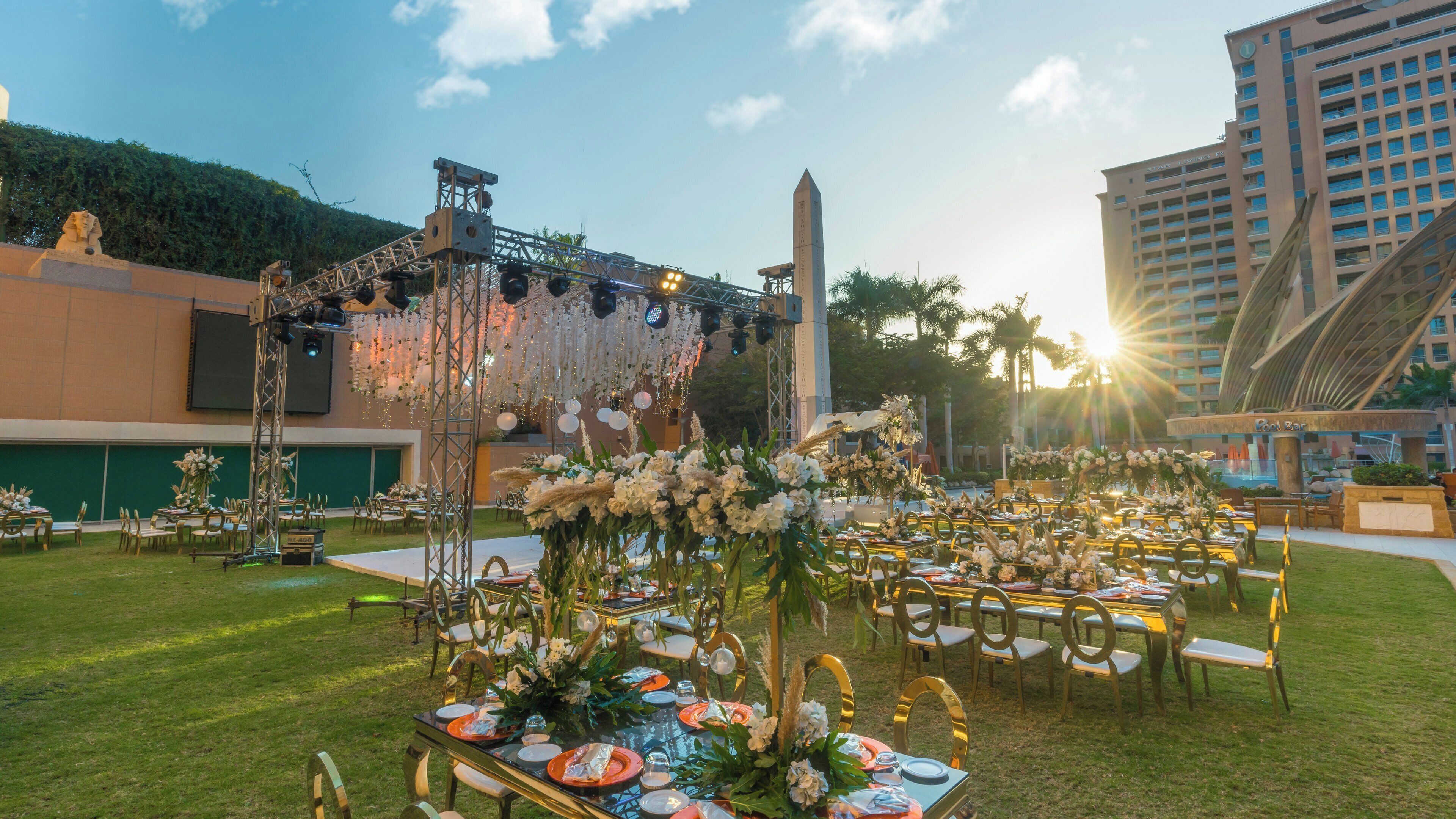 An outdoor event space with tables set for dining, a stage with lighting, and a tall monument in the background.