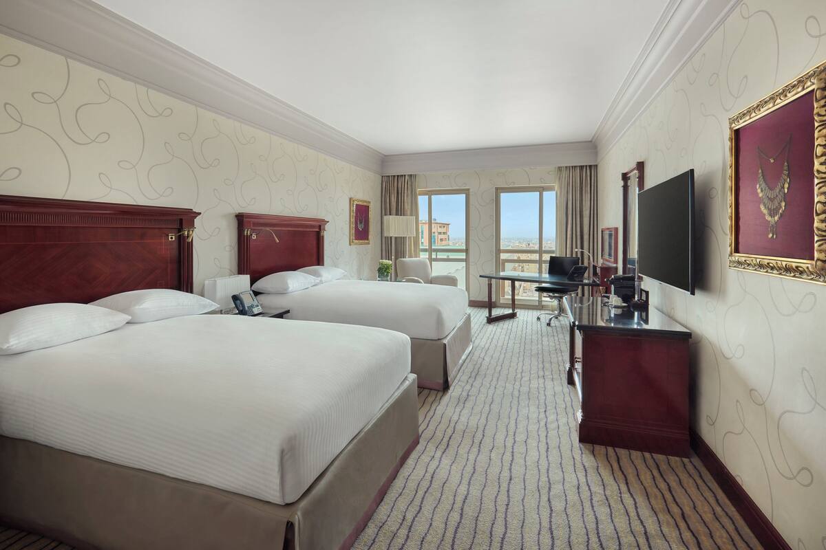 A hotel room with two beds, a desk, a TV, and a view of the city.