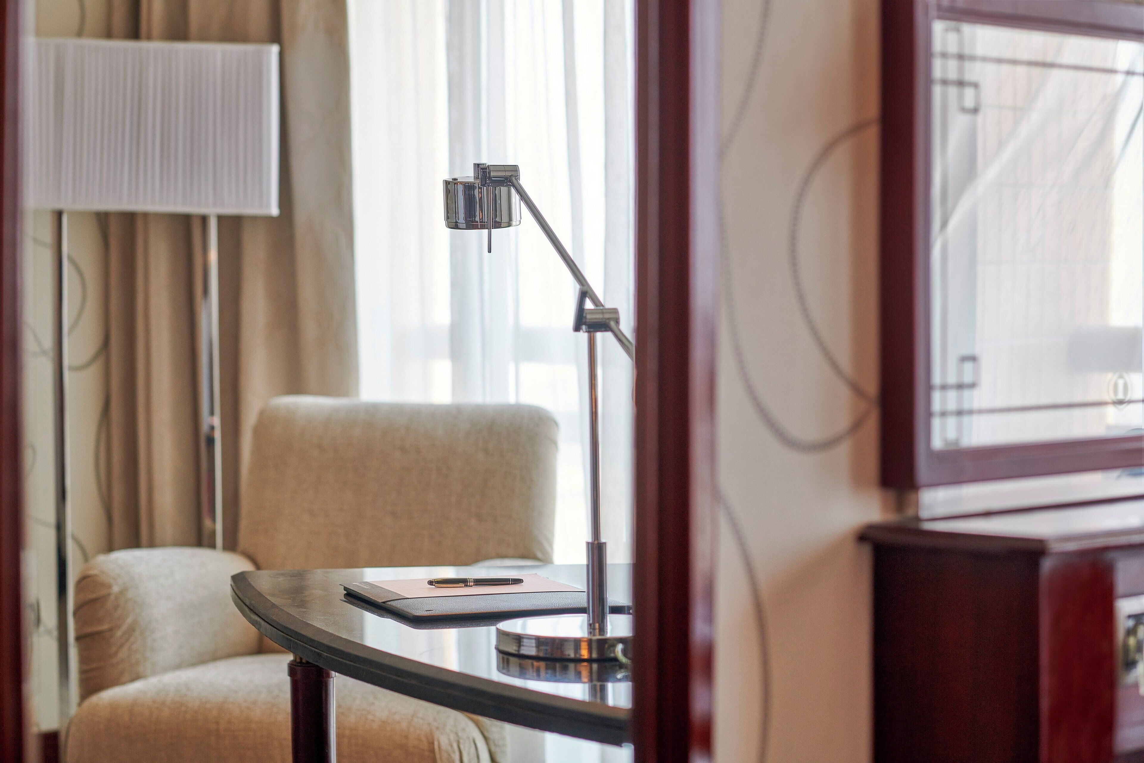 Et hotelværelse med et rundt bord, en stol, en lampe og et vindue med gardiner.