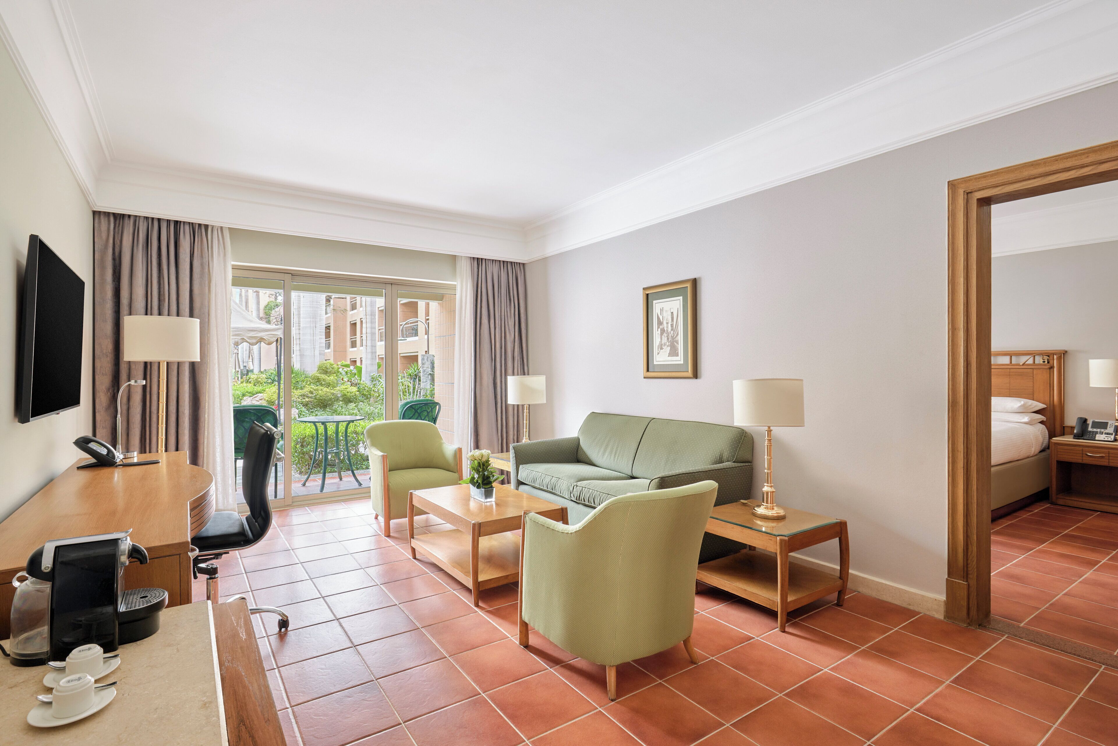 Een hotelkamer met een groene bank, een houten bureau met een koffiezetapparaat, een televisie en een balkon met uitzicht op gebouwen.