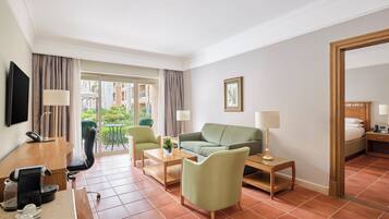 Een hotelkamer met een groene bank, een houten bureau met een koffiezetapparaat, een televisie en een balkon met uitzicht op gebouwen.