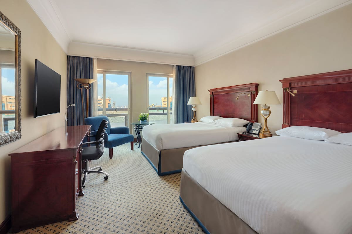 A hotel room with two beds, a desk, a chair, a TV, and a window with a view of buildings.