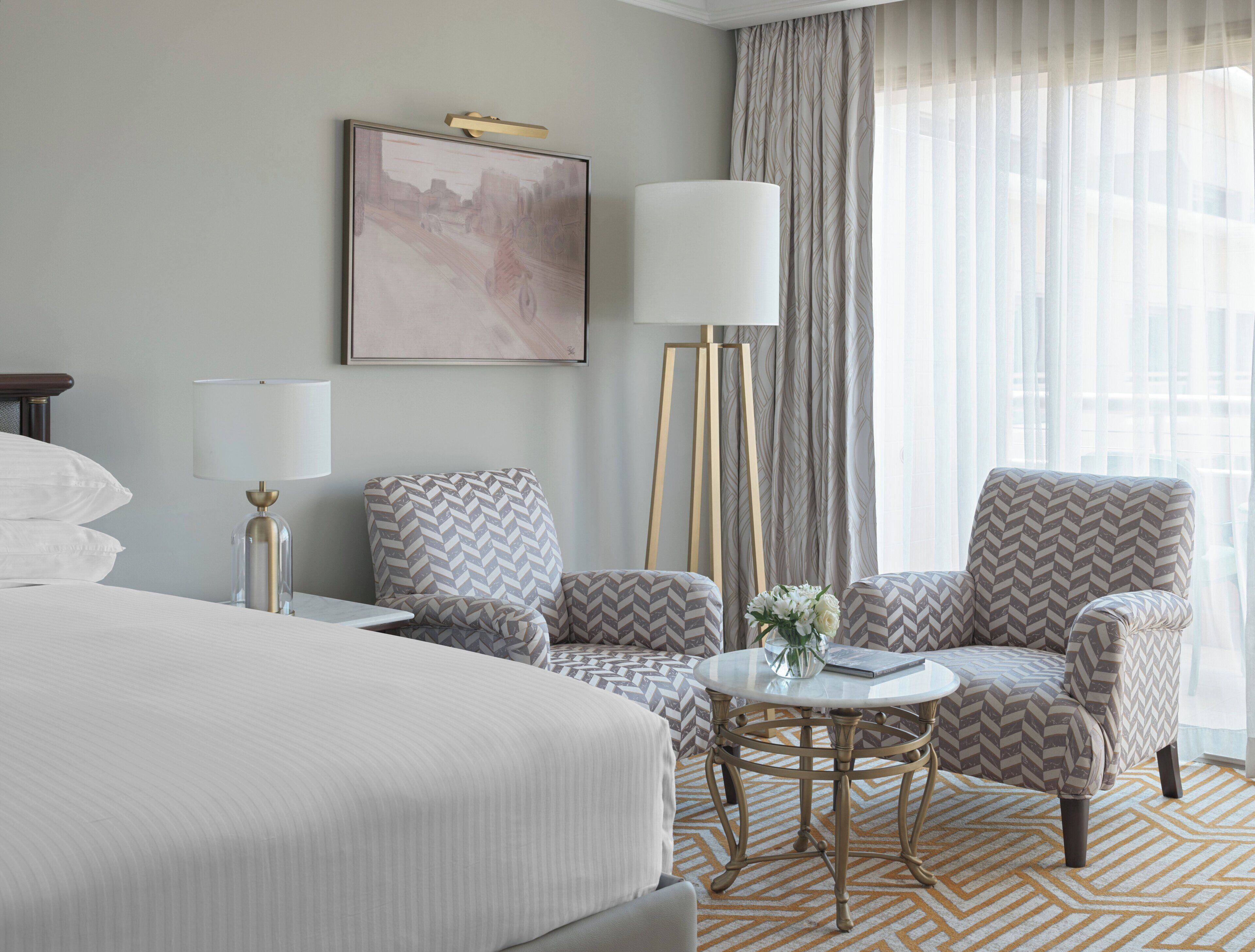 Une chambre d’hôtel avec un grand lit, deux fauteuils, une petite table sur laquelle est posé un vase de fleurs, une lampe sur pied et un tableau au mur.